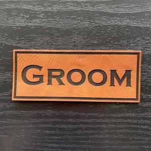 May include: A brown leather sign with the word "GROOM" embossed in black letters.