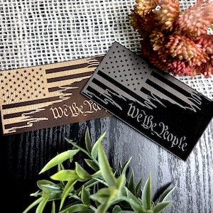 May include: Two wooden plaques with a distressed American flag design and the words "We the People" engraved on them. The plaque on the left is brown with gold stars and stripes, while the plaque on the right is black with silver stars and stripes.
