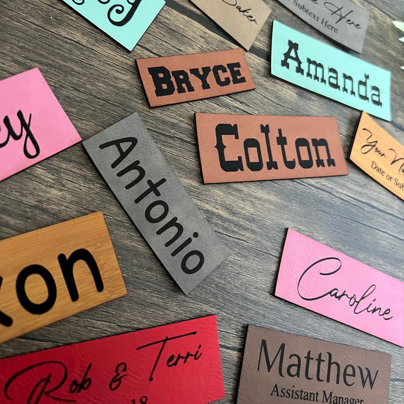 May include: A collection of personalized leather labels with various names, including Bryce, Colton, Antonio, Amanda, Caroline, and Matthew. Some labels have additional text, such as "Assistant Manager" and "Your Name, Date or Subject".