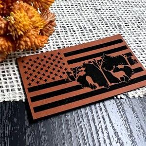 May include: A brown wooden American flag with a black silhouette of a deer and trees in the center. The flag is on a dark wood surface.