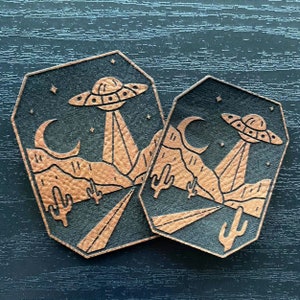 May include: Two black leather patches with a copper-colored design of a desert landscape with a UFO flying over it. The patches have a crescent moon and stars in the sky.