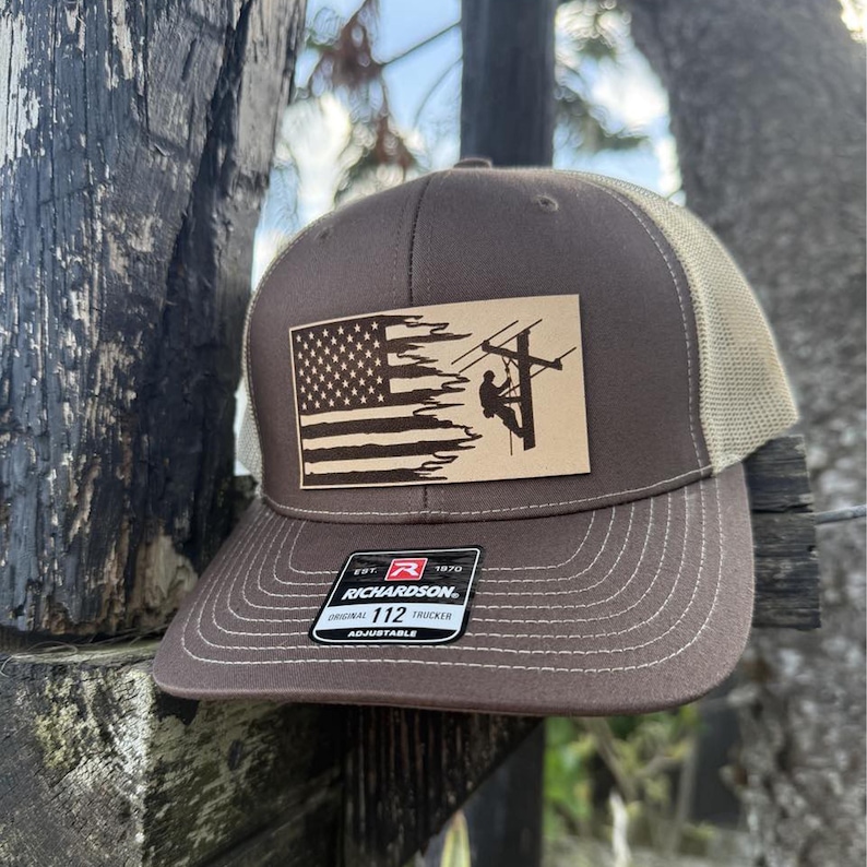 May include: Brown and tan trucker hat with an American flag and a silhouette of a lineman working on a power line. The hat has a leather patch with the text "EST. 1970 RICHARDSON. ORIGINAL TRUCKER 112 ADJUSTABLE".