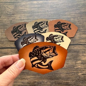 May include: A hand holds a fan of leather patches, each featuring a detailed bass fish design with an American flag motif. The patches vary in color, including shades of brown, tan, and blue, against a wooden background.