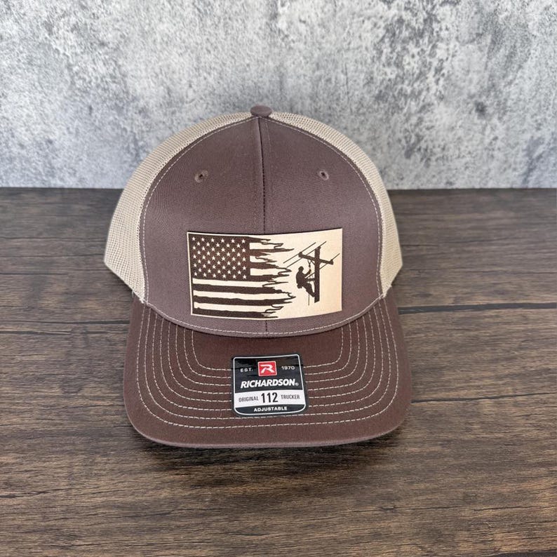 May include: Brown and tan trucker hat with a distressed American flag and a silhouette of a lineman climbing a utility pole. The hat has a Richardson 112 adjustable strap.