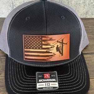 May include: Black and gray trucker hat with a brown leather patch featuring an American flag with a silhouette of a lineman working on a power line. The patch has the text "EST. 1970 RICHARDSON. ORIGINAL 112 TRUCKER ADJUSTABLE".