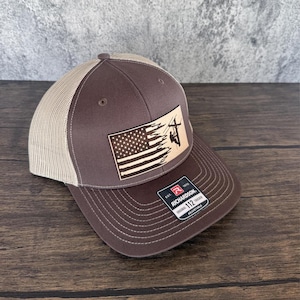 May include: A brown and tan trucker hat with a leather patch featuring an American flag and a silhouette of a lineman working on a power line. The patch is labeled "Richardson 112" and "Adjustable".