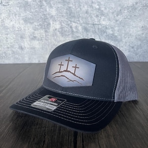 May include: A black and gray trucker hat with a black patch featuring three brown crosses on a gray mountain range. The patch is shaped like a hexagon. The hat has a black brim and a gray mesh back.