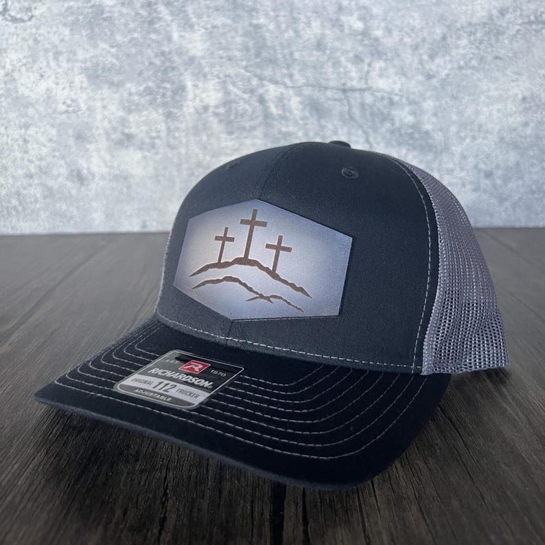May include: A black and gray trucker hat with a black patch featuring three brown crosses on a gray mountain range. The patch is shaped like a hexagon. The hat has a black brim and a gray mesh back.