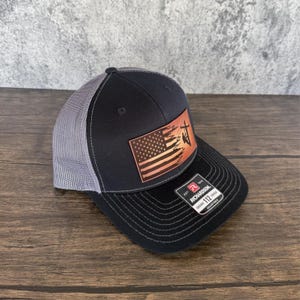 May include: A black and grey trucker hat with a leather patch featuring an American flag with a silhouette of a lineman working on a power line. The patch is brown with red, white, and blue accents. The hat has a black mesh back and a black brim. The hat has a Richardson label on the back.
