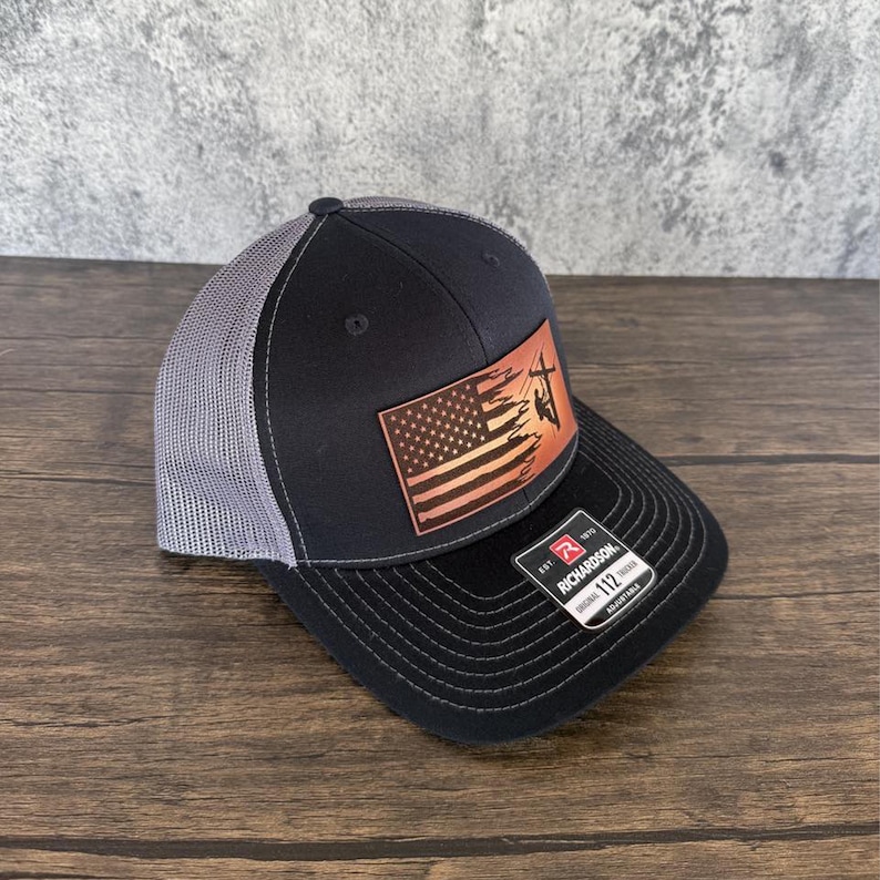 May include: A black and grey trucker hat with a leather patch featuring an American flag with a silhouette of a lineman working on a power line. The patch is brown with red, white, and blue accents. The hat has a black mesh back and a black brim. The hat has a Richardson label on the back.