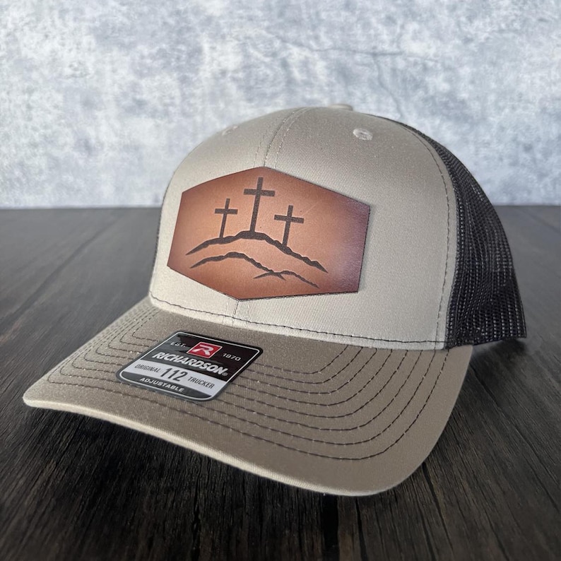 May include: A tan and black trucker hat with a brown leather patch featuring three crosses on a hill. The patch is stitched onto the front of the hat. The hat is adjustable and has a Richardson 112 Trucker label.