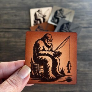 May include: A brown leather coaster with a black silhouette of a Bigfoot sitting on a log and holding a fishing rod with a fish on the line. The background is a light orange color.