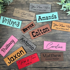 May include: A collection of personalized leather name tags in various colors, including brown, red, pink, orange, gray, and blue. The tags feature names such as Brittney, Jaxon, Colton, Amanda, Bryce, and Matthew. Some tags also include additional text, such as "Assistant Manager" and "John 3:16".