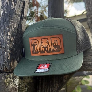May include: Green flat bill snapback hat with a brown leather patch that says "DAD" with deer and archer graphics. The hat has a mesh back and a flat bill. The hat is a Richardson brand.