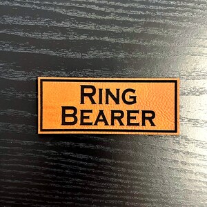 May include: A brown leather tag with black text that reads "RING BEARER".