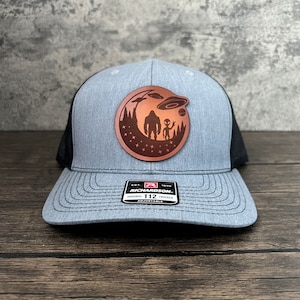 May include: A grey and black trucker hat with a brown leather patch featuring a silhouette of Bigfoot and an alien with a UFO in the background. The patch has a starry night sky design. The hat has the text "Richardson 112 Original Trucker Adjustable" on the front.
