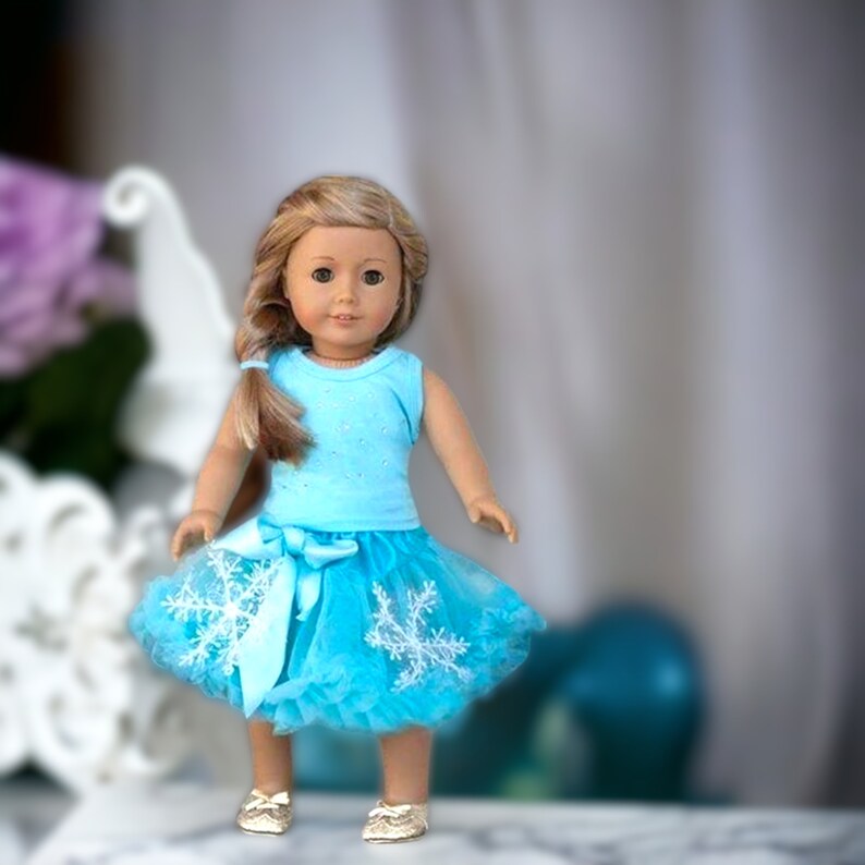 Puede incluir: Una mu&ntilde;eca con un top azul sin mangas con un dise&ntilde;o brillante y una falda de tul azul con detalles de copo de nieve blanco. La mu&ntilde;eca tiene el pelo largo y casta&ntilde;o y los ojos azules.