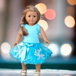 Puede incluir: Una mu&ntilde;eca con un top azul claro sin mangas con un dise&ntilde;o brillante y una falda de tul a juego con adornos de copo de nieve. La mu&ntilde;eca tiene el pelo largo y rubio y los ojos marrones.