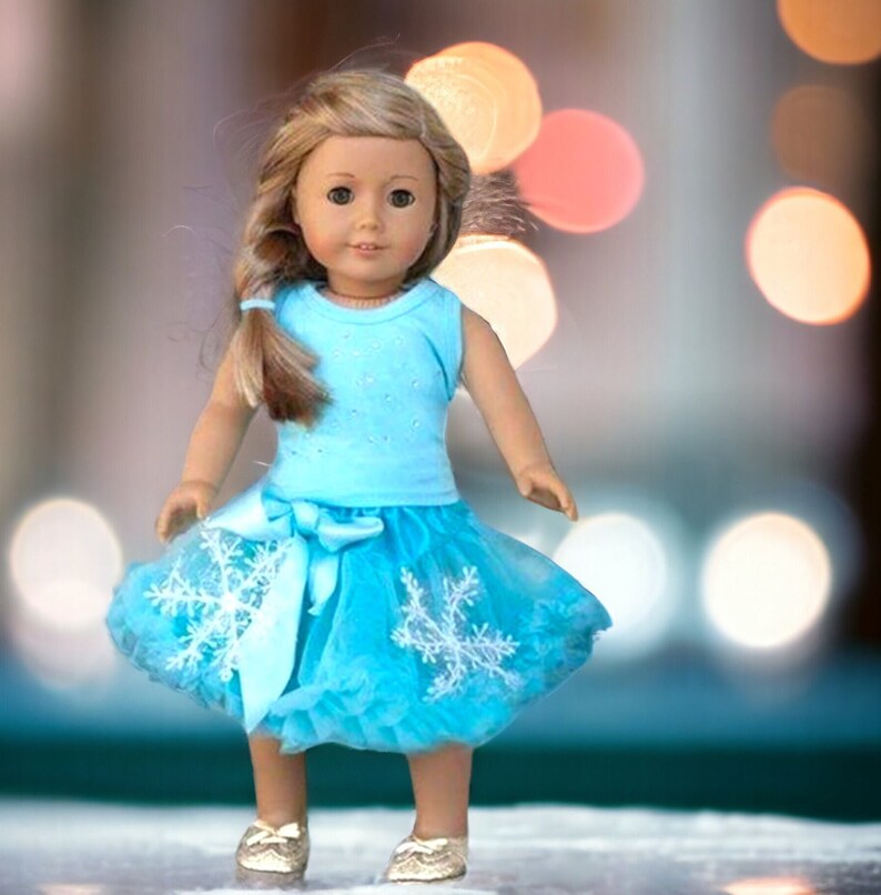 Puede incluir: Una mu&ntilde;eca con un top azul claro sin mangas con un dise&ntilde;o brillante y una falda de tul a juego con adornos de copo de nieve. La mu&ntilde;eca tiene el pelo largo y rubio y los ojos marrones.