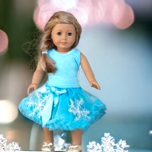 Puede incluir: Una mu&ntilde;eca con un vestido azul claro sin mangas con un estampado de copo de nieve blanco y una falda de tul azul a juego. La mu&ntilde;eca tiene el pelo largo y rubio y los ojos azules.
