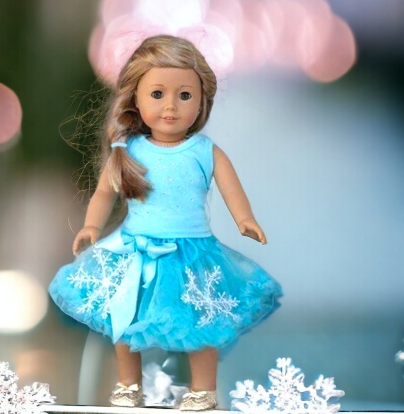 Puede incluir: Una mu&ntilde;eca con un vestido azul claro sin mangas con un estampado de copo de nieve blanco y una falda de tul azul a juego. La mu&ntilde;eca tiene el pelo largo y rubio y los ojos azules.