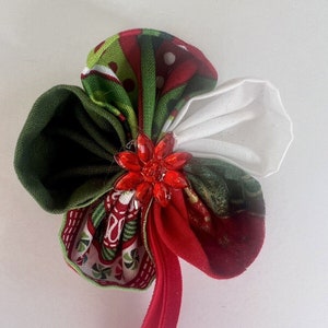 Peut inclure: Une fleur en tissu faite à la main avec un centre en bijou rouge et un élastique rouge. Les pétales de la fleur sont un mélange de tissus rouges, verts et blancs, certains à motifs. Un accessoire festif.