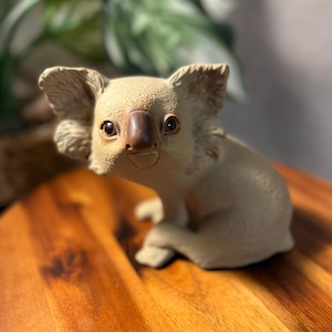 May include: A beige koala figurine with a brown nose and dark eyes. The koala is sitting on a wooden surface. The figurine has detailed features and a friendly expression.