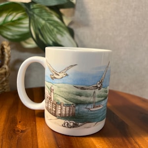 May include: White ceramic mug featuring a coastal scene with flying seagulls, a sailboat, and a building with a tower. The mug has a curved handle and is set against a wooden surface.