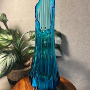 May include: A tall, turquoise-blue glass vase with a fluted design. The vase has a unique, asymmetrical top and a rounded base. The glass is transparent, allowing light to pass through, and is approximately 12 inches tall.