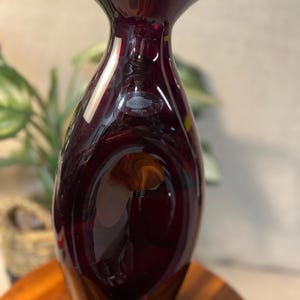 May include: A deep burgundy glass vase with a smooth, rounded, teardrop-shaped body and a flared rim. The vase features a clear, oval-shaped sticker on the front surface with text that reads Made in USA, reflecting light on its polished surface.