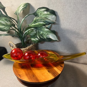 May include: A decorative glass dish with a yellow stem and a clear, elongated bowl. Three red glass spheres rest inside. The dish sits on a wooden stand, with a potted plant in the background.