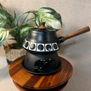 May include: A matte black fondue pot with a wooden handle and a matching wooden lid knob. The pot features a decorative band of white squares containing black circles. It sits on a black metal base stand that houses a small burner underneath.