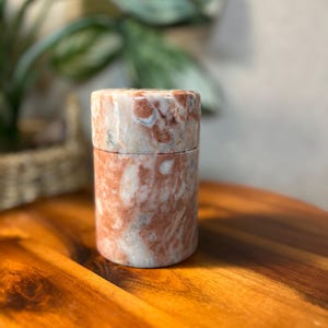 May include: A cylindrical container with a lid, crafted from a stone with a marbled pattern of red, pink, and cream hues. The container sits on a wooden surface, with a blurred background of greenery.