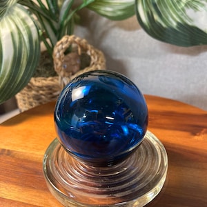 May include: A deep blue glass sphere sits atop a clear, circular glass base with concentric rings. The sphere reflects light, creating a vibrant, glossy appearance. The base is on a wooden surface, with green and white leafy plants in the background.