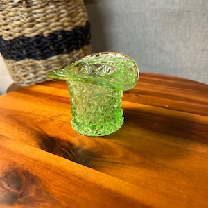 May include: A small, green glass top hat-shaped container with a detailed starburst pattern. The container is sitting on a wooden surface. A woven basket is in the background.