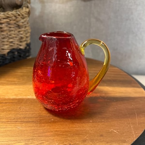 May include: A small, red glass creamer with a yellow handle. The creamer has a crackle finish and a rounded shape. The handle is curved and attached to the side of the creamer. The creamer is sitting on a wooden surface.