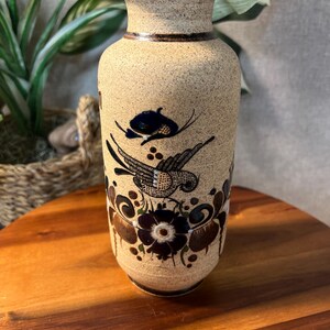 May include: A beige ceramic vase with a textured surface and a dark brown rim. The vase features a hand-painted design of a bird and floral motifs in dark blue and brown. The vase is displayed on a wooden surface.