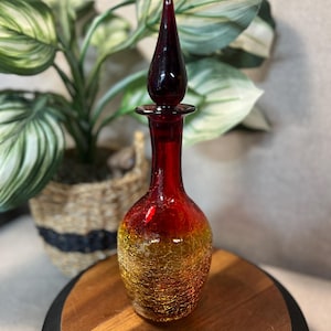 May include: A decorative glass bottle with a red stopper and a crackle-effect body that transitions from red to gold. The bottle has a long neck and a bulbous base. The background includes a potted plant.