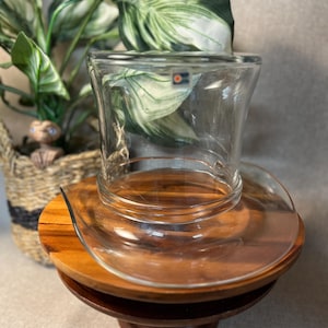 May include: A clear glass top hat shaped vessel with a wide brim and a cylindrical crown. The glass features a small square sticker with an orange circle logo near the top rim. The object is positioned on a round wooden pedestal base.
