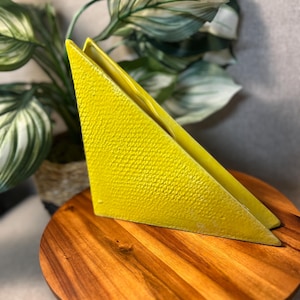 May include: A bright yellow, triangular napkin holder with a textured surface. The holder is displayed on a round, wooden stand. Green and white leafy plants are in the background.