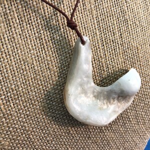 May include: A white, organic, shell-shaped pendant hangs from a brown leather cord. The pendant is smooth and has a natural, irregular shape.