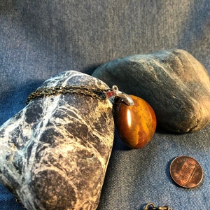 Puede incluir: Un colgante de piedra marrón y naranja en una cadena de plata. El colgante tiene forma de lágrima y tiene un acabado liso y pulido. La cadena está hecha de pequeñas piezas de metal enlazadas.