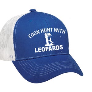 May include: A blue and white trucker hat with the text "COON HUNT WITH LEOPARDS" in white. The hat has a mesh back and a curved brim. The text is above a graphic of a tree and two figures.