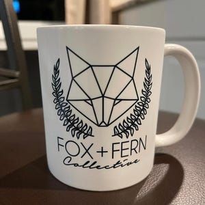 May include: White ceramic mug featuring a geometric fox head design in black, framed by laurel branches. The text "FOX + FERN Collective" is printed below the fox illustration. The mug has a curved handle.