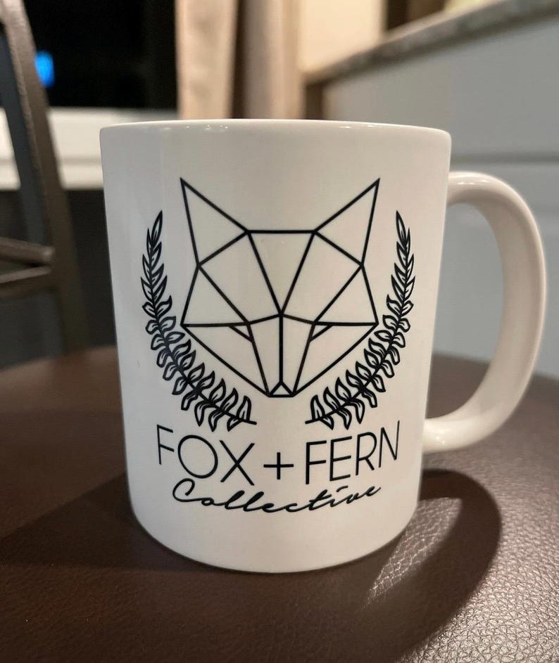 May include: White ceramic mug featuring a geometric fox head design in black, framed by laurel branches. The text "FOX + FERN Collective" is printed below the fox illustration. The mug has a curved handle.