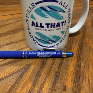 May include: A white mug with the text "ALL THAT! MOVING AND MORE" and a blue and teal design. A blue pen with silver accents and the text "ALL THAT MOVING JACKSONVILLE LLC" is next to the mug.