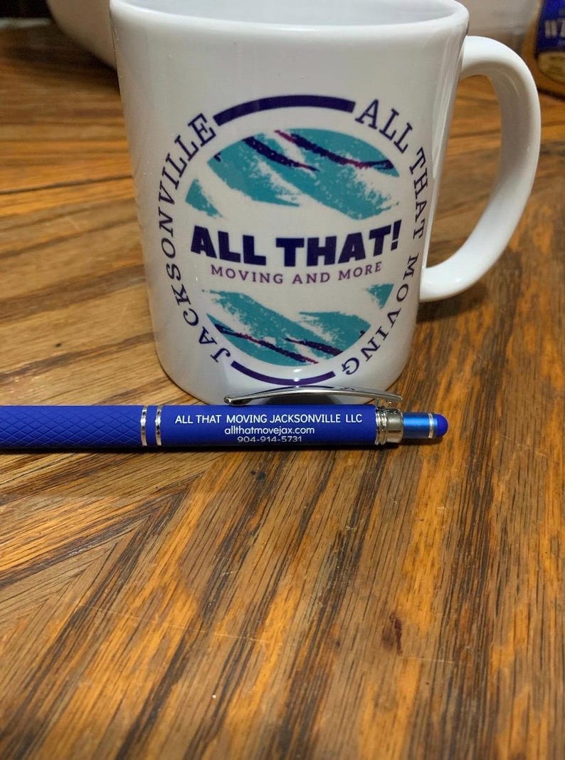 May include: A white mug with the text "ALL THAT! MOVING AND MORE" and a blue and teal design. A blue pen with silver accents and the text "ALL THAT MOVING JACKSONVILLE LLC" is next to the mug.