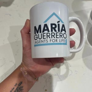 May include: White ceramic mug with the text "MARIA GUERRERO AGENTS FOR LIFE" in black and a blue house logo. The mug has a curved handle and is held in a hand.
