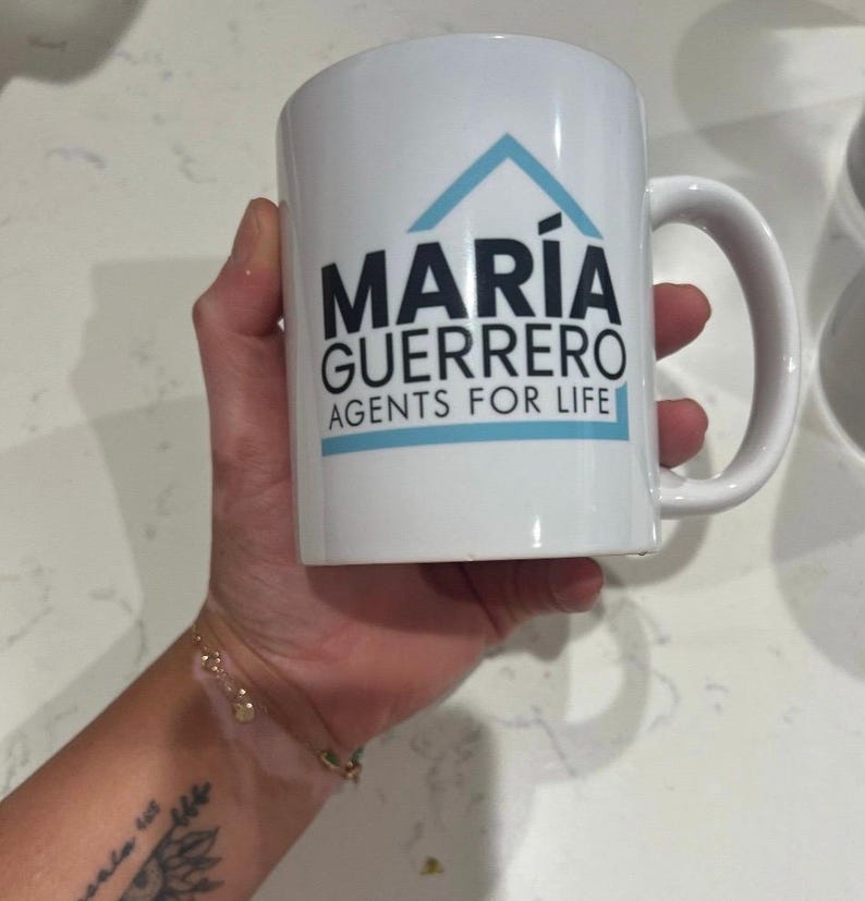 May include: White ceramic mug with the text "MARIA GUERRERO AGENTS FOR LIFE" in black and a blue house logo. The mug has a curved handle and is held in a hand.