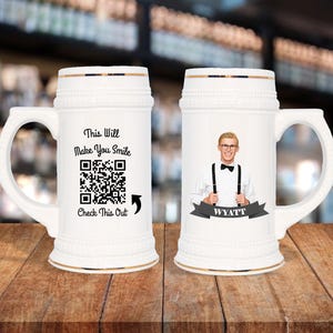 May include: White ceramic beer stein with a handle, featuring gold trim and a decorative top. One stein has a QR code and the text "This Will Make You Smile, Check This Out". The other stein has a photo of a man and the name "Wyatt".